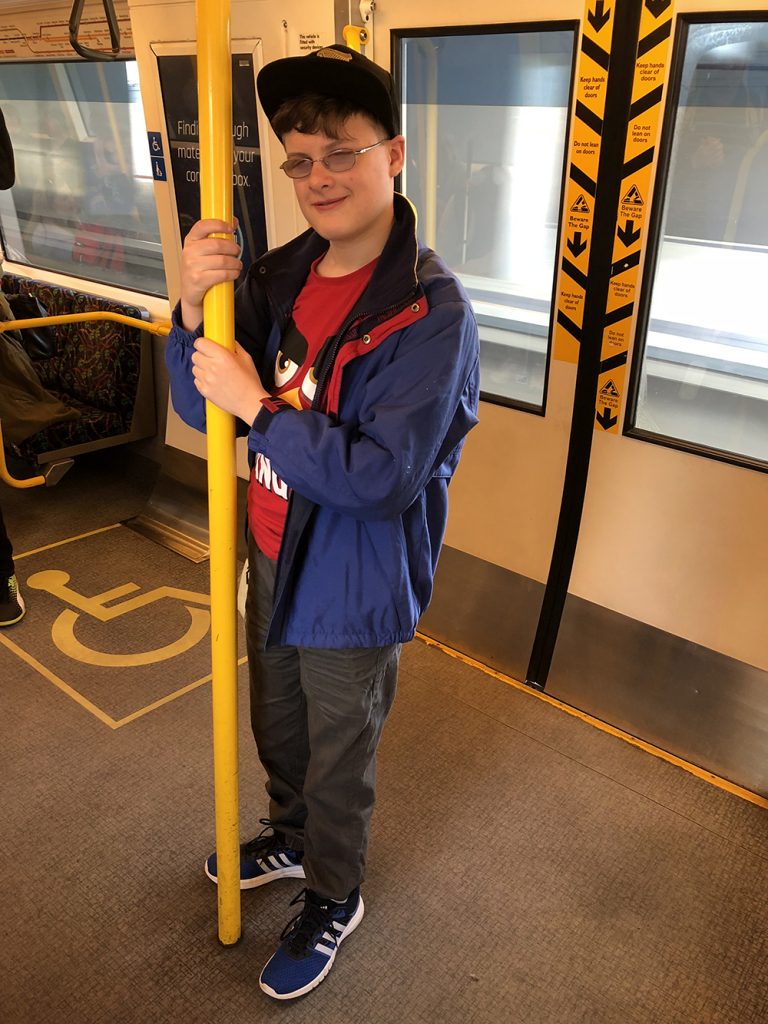 Rishi stands holding on to a pole in a train carriage. He is wearing a red shirt, and purple jacket.