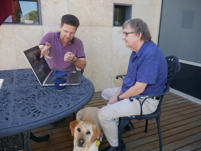 Therapist David Vosnacos sitting down with a client looking at a laptop with a Guide Dog sitting next to the client.
