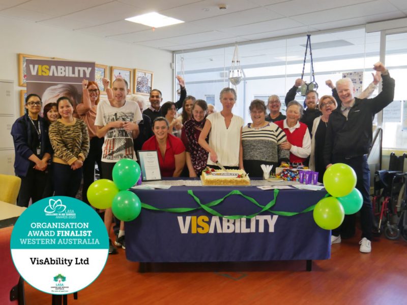Staff and clients gathered around a table with our CEO Elizabeth Barnes cutting into a large carrot cake. VisAbility logo is pictured in the corner.