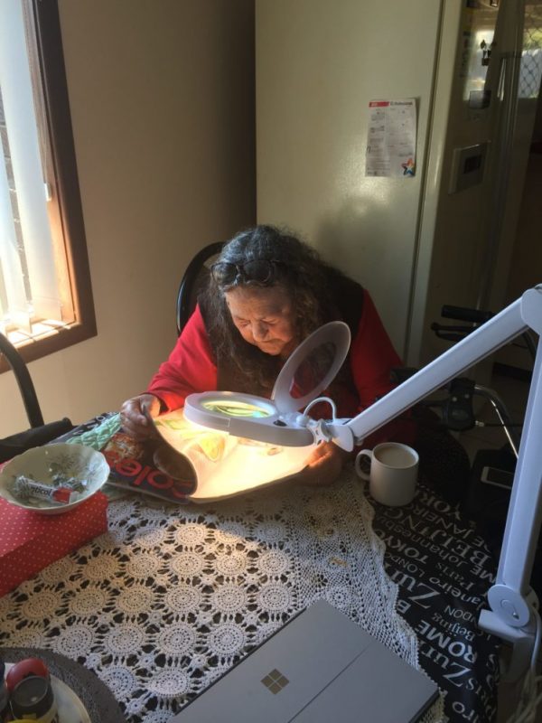 Kaye uses a light screen to read a daily paper. She's sitting around a table. Regional therapist Jack Beer visits her at her farm outside of Geraldton