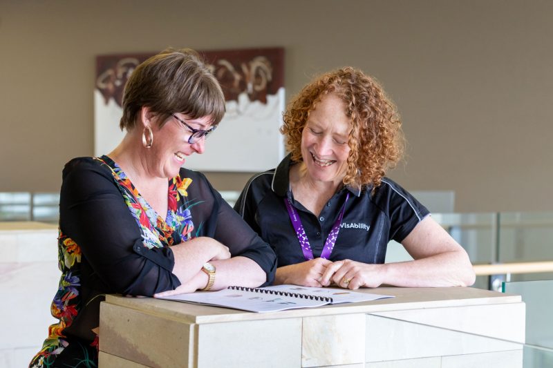 VisAbility staff member in uniform standing next to client, talking and pointing to booklet in front of them