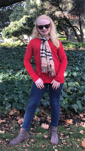 Image of Karin standing in a lush green garden, wearing a red top, a scarf, and blue jeans.