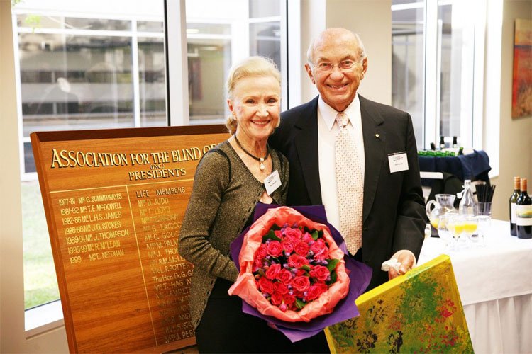 Jean and Stan Perron holding flowers and a gift in front of the Association for the Blind Life Members board