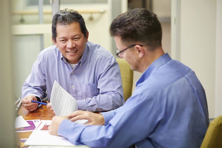Image of two men sitting at a desk looking at file paper.