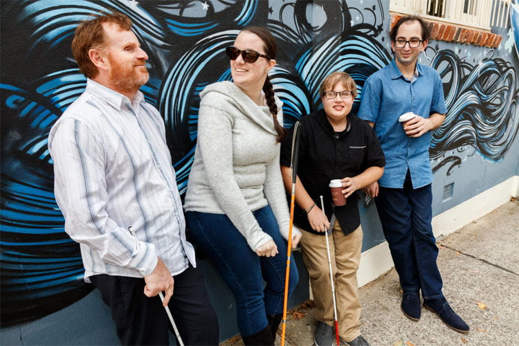 Image of a VisAbility team member with three young adults, leaning against a coloured mural.