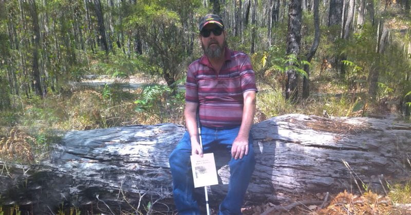 Phil sits on a log in a forest. He is holding his white cane