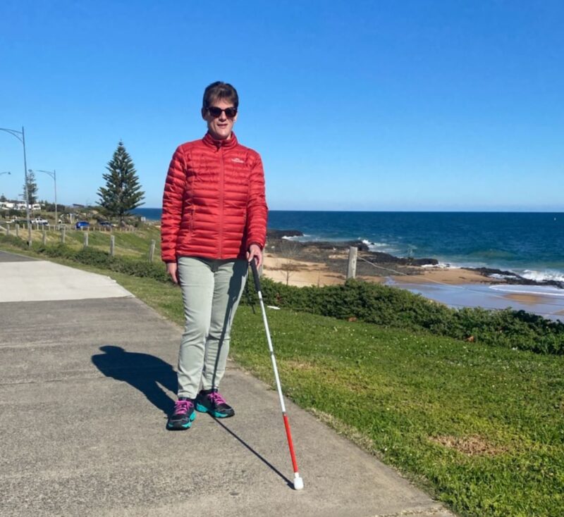 Image shows Nicole with her white cane walking along a footpath close to the sea