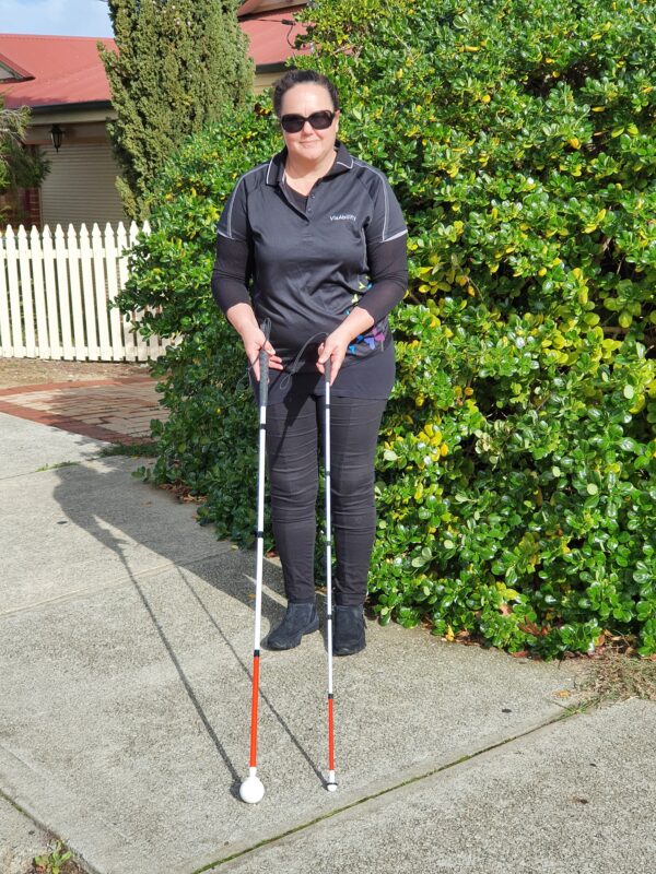 Jodie stands in front of a tree on a path showing the long cane and a fold away cane