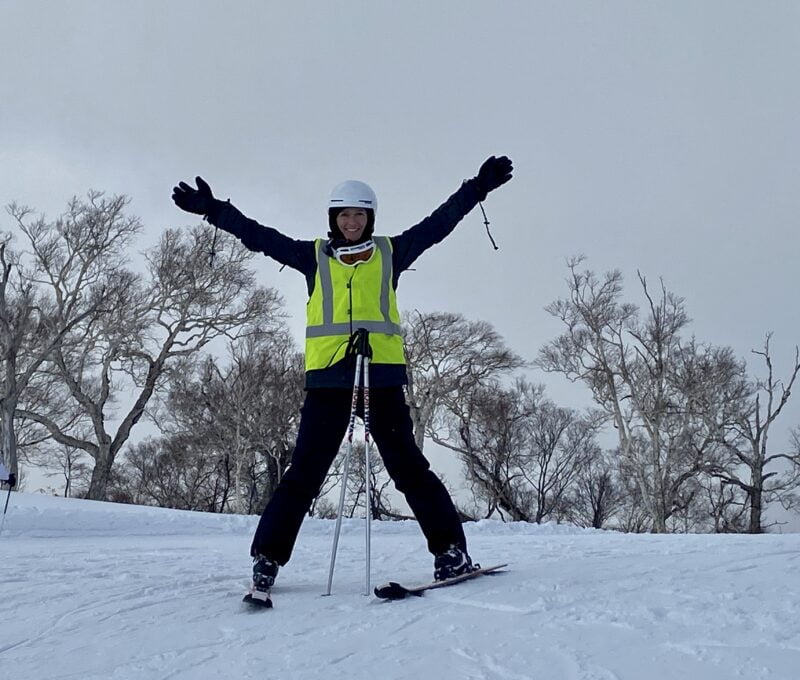 Melanie stands on snow slope with arms outstretched in flourescent jacket