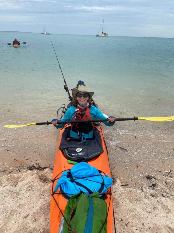 Melanie sits in an orange kayak - half in sea half in water 