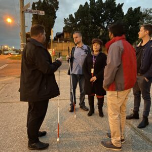 A group of people holding white canes stand in the street talking to O&M instructor