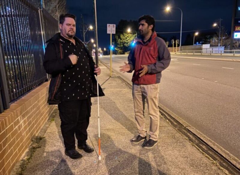 Man with white cane stands on pavement alongside O&M instructor who's explaining more about the cane