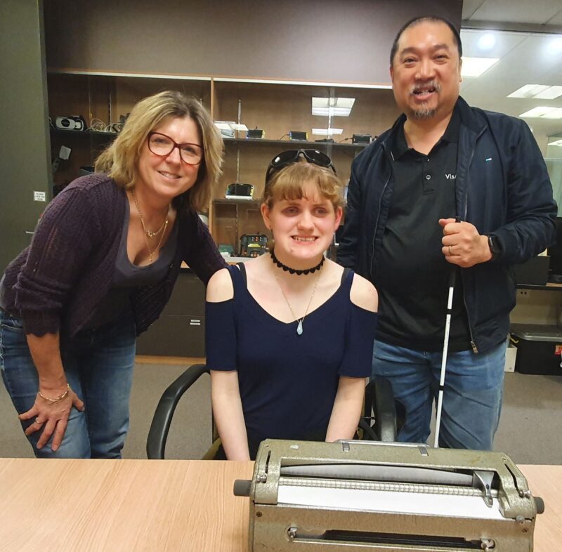 Mel stands next to Tayla and Manny. A Perkins Brailler is on the table in front of them in the assistive technology room.