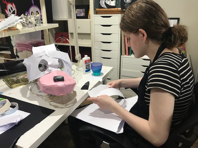 Tayla sits at a crafting table with her mask and in her hands she holds sheets of card