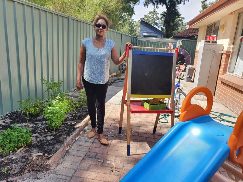 Image shows saba standing in her garden next to a chalk board with a child's slide to one side and garden border to one side 