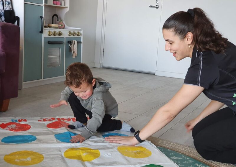 Mateo is leaning over a Twister mat and placing his hand on a blue circle following instruction from Speech Pathologist Helena
