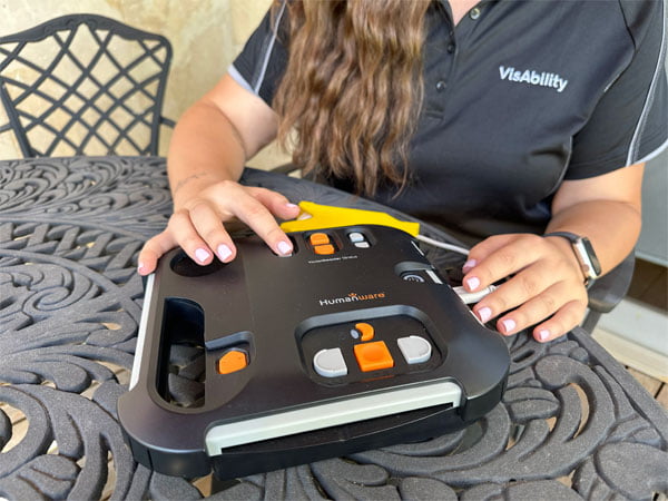Woman sits in front of the audio book player which is laid out on a table. Hand is poised over a large orange button.