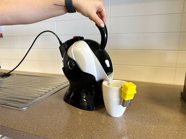 A hand is on the handle of a tilting kettle tipped slightly with water pouring into a mug.