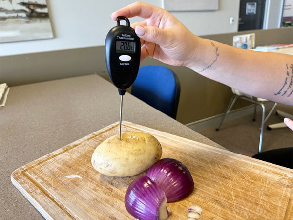 Person holds thermometer into a potato which is on a chopping board next to a cut onion.