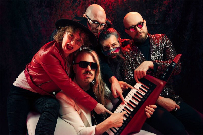 Simone and four other members of the band gather around a small organ. They are all trying to play the organ and are in a fun pose, smiling and giggling