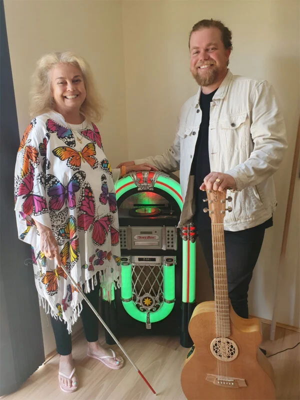 Ricky and his mum Anne stand net to each other around a jukebox. Anne is holding her white cane and Ricky holds his guitar.