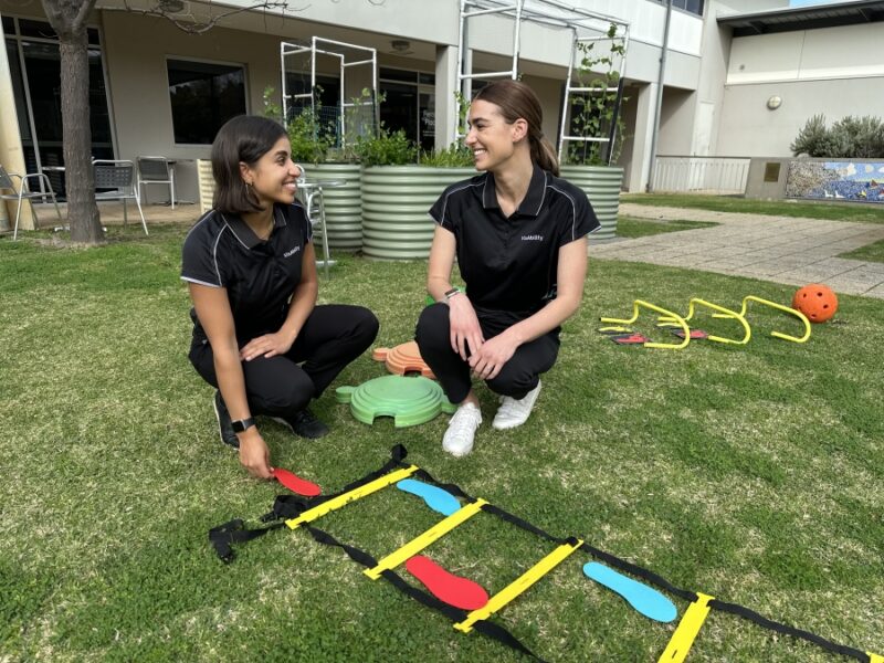 Our two physiotherapists Emma and Yas kneel next to each other on grass with play equipment