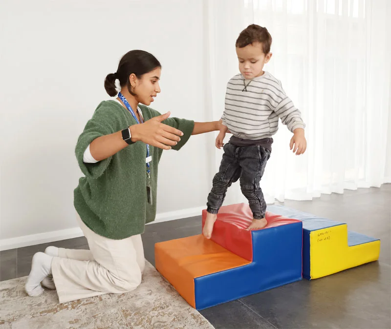 Physiotherapist Yasmin works with a client supporting him to balance. Child walks over a soft step. 