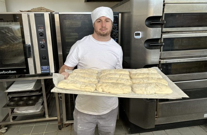 Rhys stands in front of an oven holding a large tray of dough ready to be baked into French baguettes.