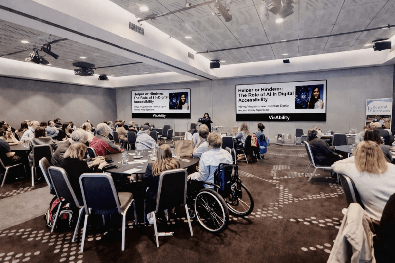 A wide-angle shot of the conference room, showing Vithya speaking to a room full of people.